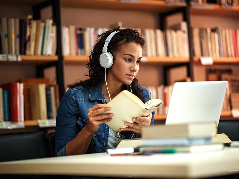 A grad student consults a book while taking an online course on her laptop.